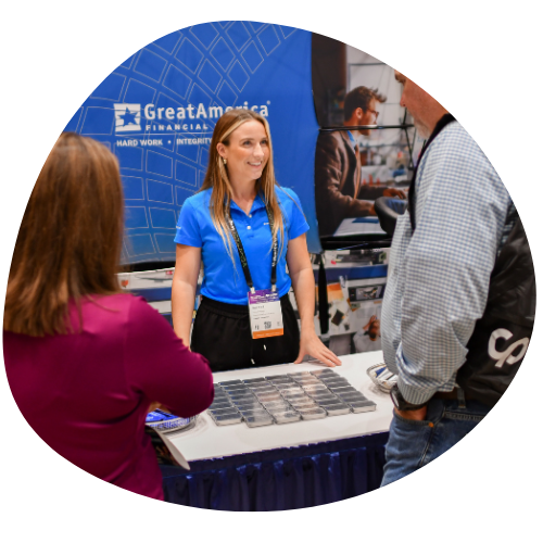 A photo of a woman standing behind an exhibitor table, smiling and chatting with two people who are on the opposite side of the table. 