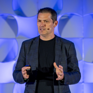 A photo of a man wearing a dark blue jacket speaking onstage