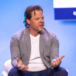 A photo of a man with a light grey jacket sitting and speaking onstage