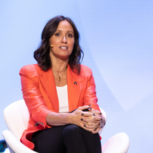 A photo of a woman with medium-length brown hair and a red blazer sitting and speaking onstage